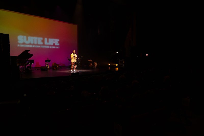 A performer in a patterned outfit stands on a stage with a large screen displaying "SUITE LIFE" in gradient colors.