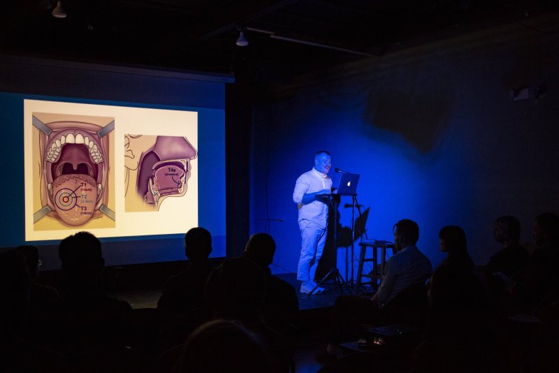 Presentation in dark room. Speaker stands at laptop under blue light, beside screen showing anatomical diagrams. Silhouetted audience in foreground.