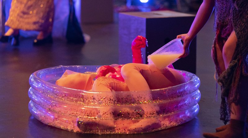 Performance art featuring a person in a glittery inflatable pool, wearing peach-colored clothing. Another person pours a yellow liquid from a clear container onto them. Red inflatable objects are also in the pool. The scene is lit with purple and pin
