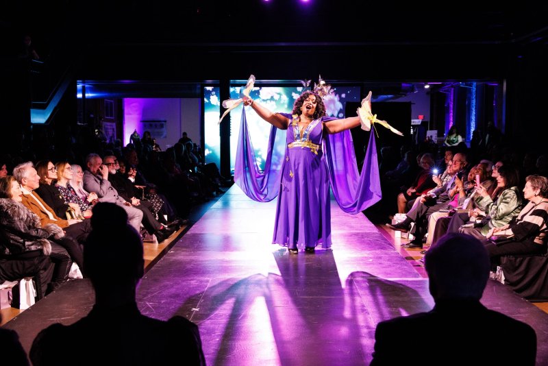 Drag queen in a flowing purple gown with gold accents and a feathered headpiece, arms outstretched on a runway, audience seated on both sides.