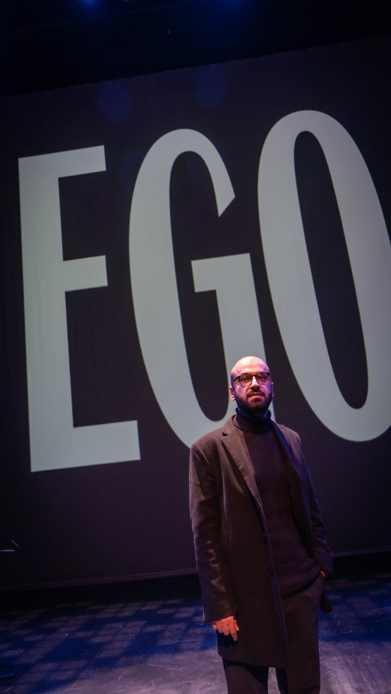 A man with glasses and a dark coat stands before a large screen displaying the word "EGO" in white sans-serif font.