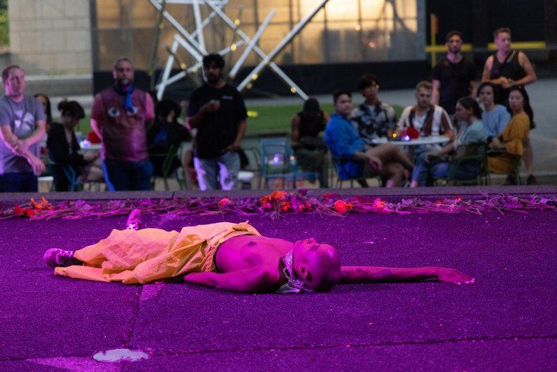 Performance art piece with a person lying on asphalt under purple light, wearing a yellow garment and jewelry. A line of flowers separates the figure from a blurred audience in the background.