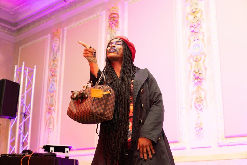 Performer with blue face paint, red beret, and long braids points upward, holding a patterned bag. Stage lighting is pink and purple. Ornate wall details are visible in the background.