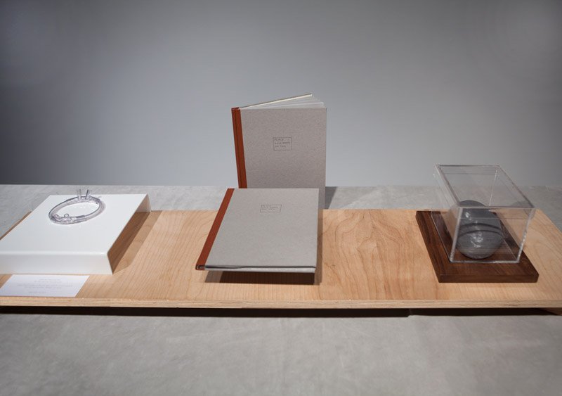 A light brown wooden tray displaying two gray books with orange spines, a clear bracelet on a white stand, and a gray object in a clear box.