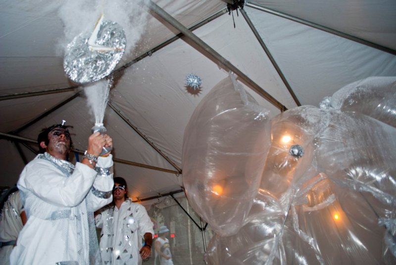 Man in white and silver costume sprays a bottle, creating a misty cloud around a silver disco ball under a tent.