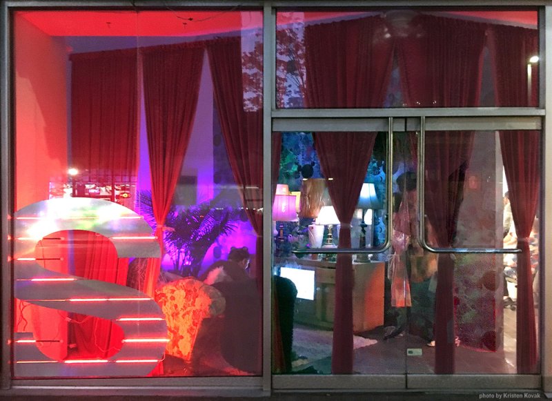 Storefront window display with red curtains, neon "S" sign, purple lighting, and lamps illuminating a room.