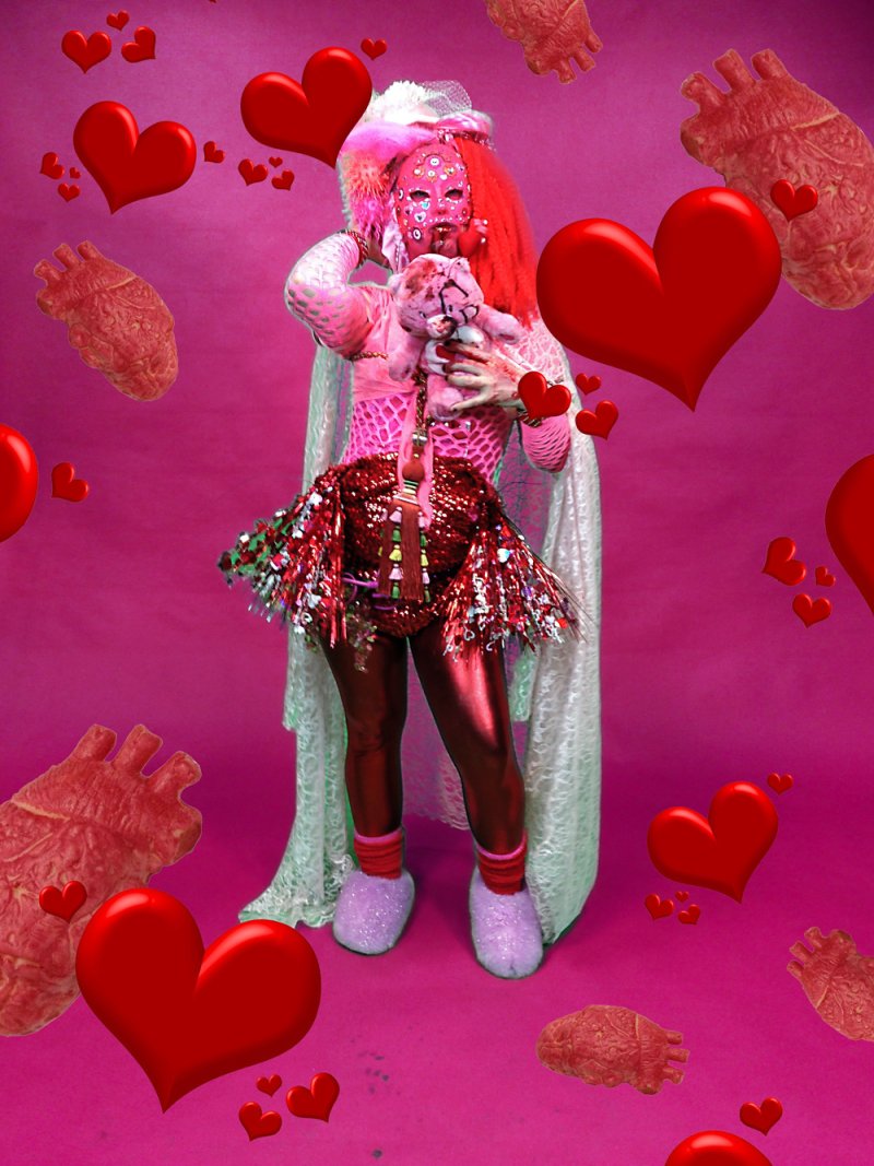Figure in elaborate pink costume with red wig and patterned mask, holding a stuffed animal. Red hearts and anatomical hearts float against a pink backdrop. The figure wears a sequined skirt and iridescent cape.
