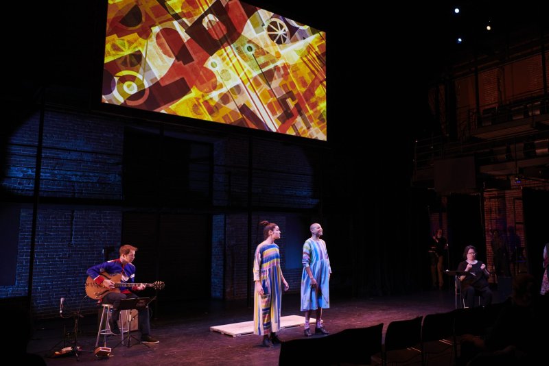 Stage performance with two singers in striped dresses, guitarist, and abstract projection. Brick wall backdrop. Colors include yellow, red, blue.