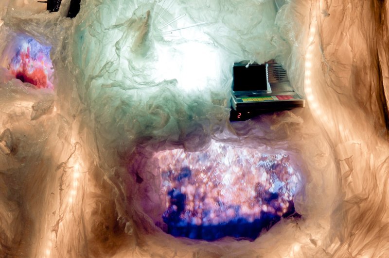 Crumpled white plastic installation with glowing pink, purple, and blue projections, and a black laptop on a shelf.