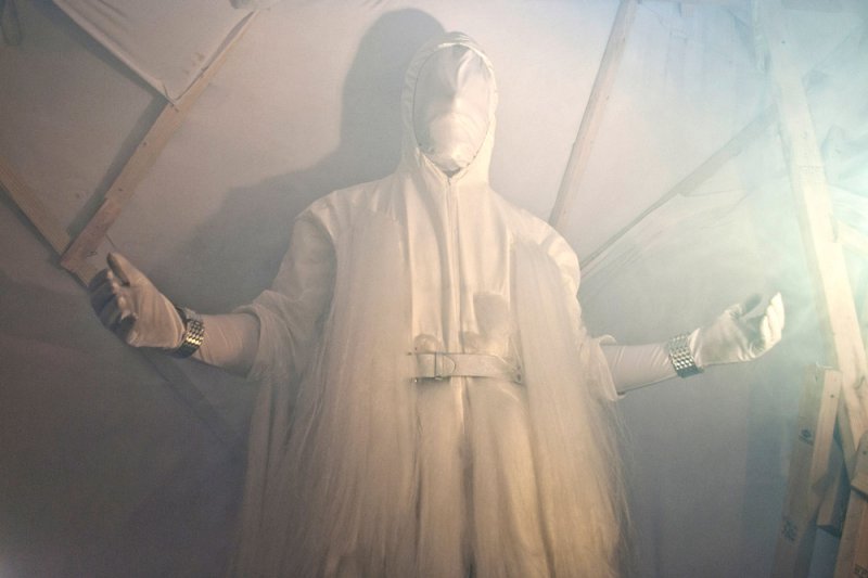 Figure in a white hooded suit with a blank face, long white hair, and outstretched gloved hands, standing in a hazy room.