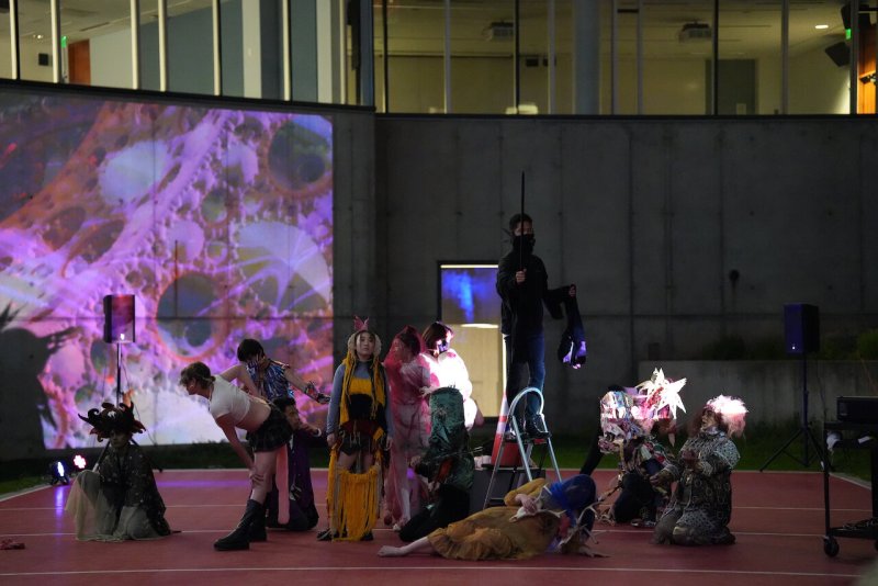 Performance art with costumed figures on red court, projected with pink and purple abstract patterns on a wall.