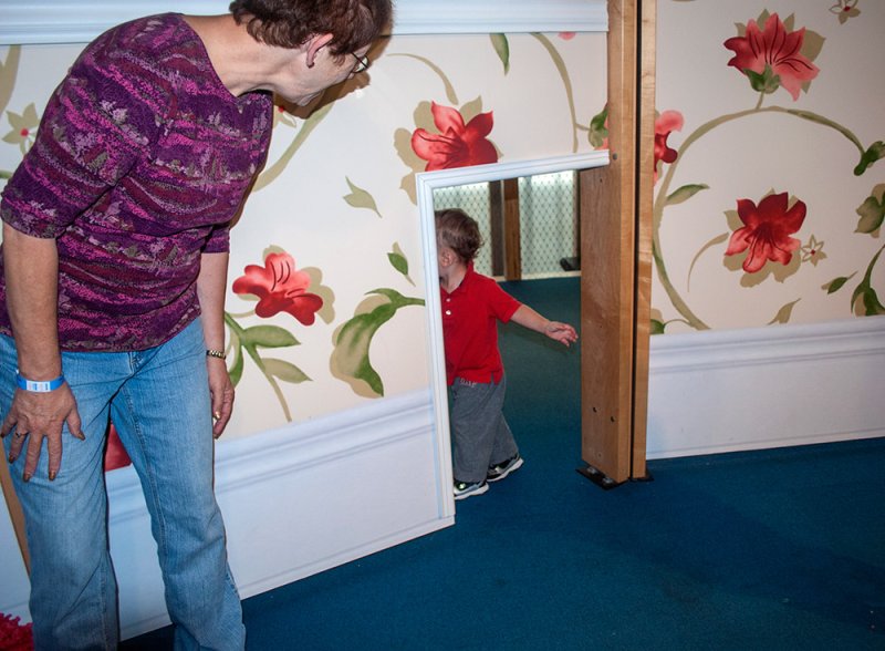 Woman in purple shirt and jeans peers into a mirror, reflecting a toddler in a red shirt walking away.