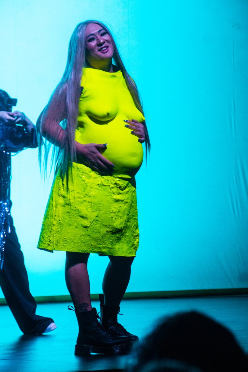 Performer with long blonde hair, wearing a neon yellow top with breasts and a matching textured skirt, smiling.