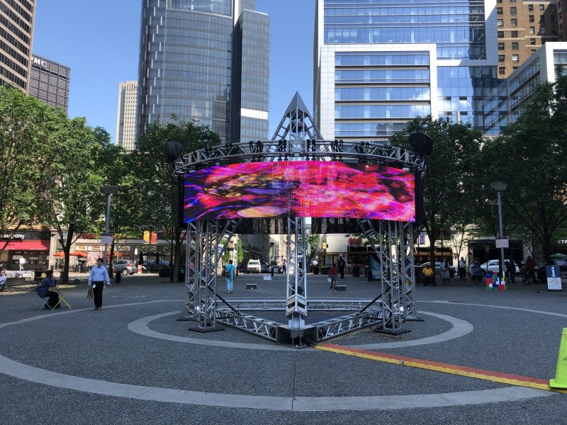 Outdoor installation: metal frame supports a vibrant LED screen with abstract pink, purple, and blue patterns. Urban setting.