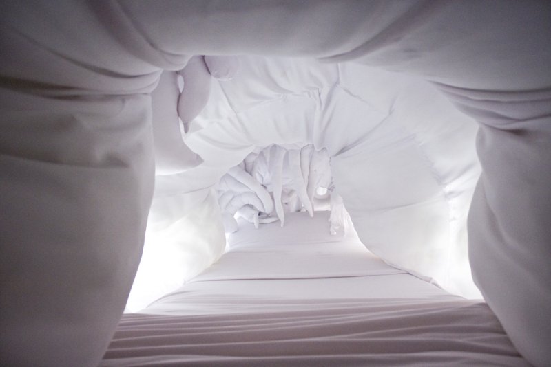 Looking up into a narrow, white fabric tunnel with draped and ruffled textures, illuminated by soft light.