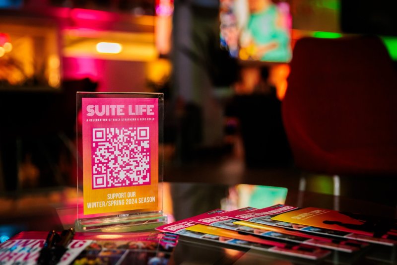 A pink and orange "Suite Life" QR code display on a table, surrounded by brochures, with blurred neon lights in the background.