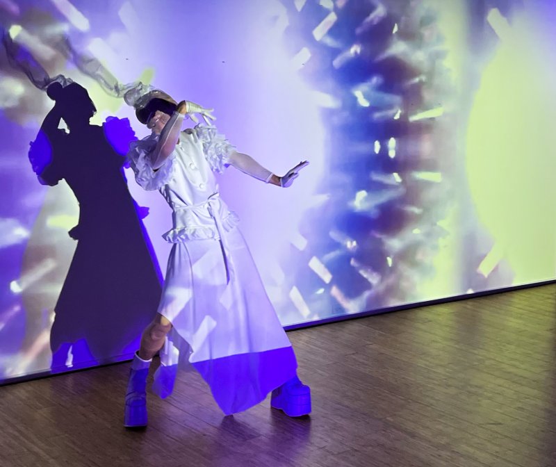 Performer in a white avant-garde outfit, platform shoes, and mask, posing against a purple and green projected digital backdrop.