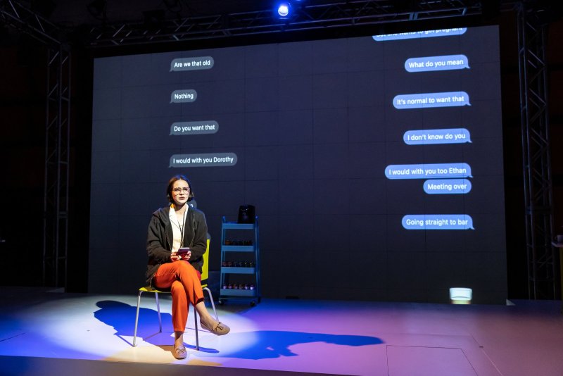 A stage scene with a woman seated on a yellow chair, holding a phone. Behind her, a screen displays text messages in speech bubbles. The stage is lit with blue light.