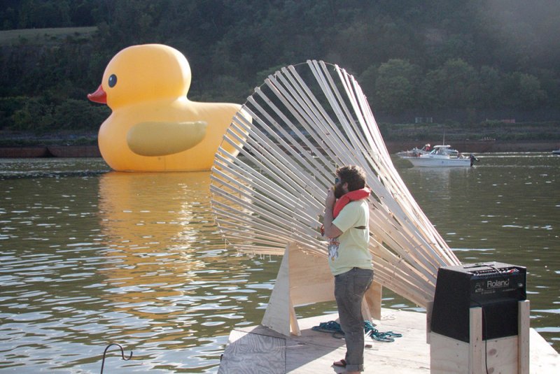 Outdoor art installation on water featuring a large inflatable yellow duck and a wooden fan-like structure. A man in a life vest stands on a raft, with a boat visible in the background.