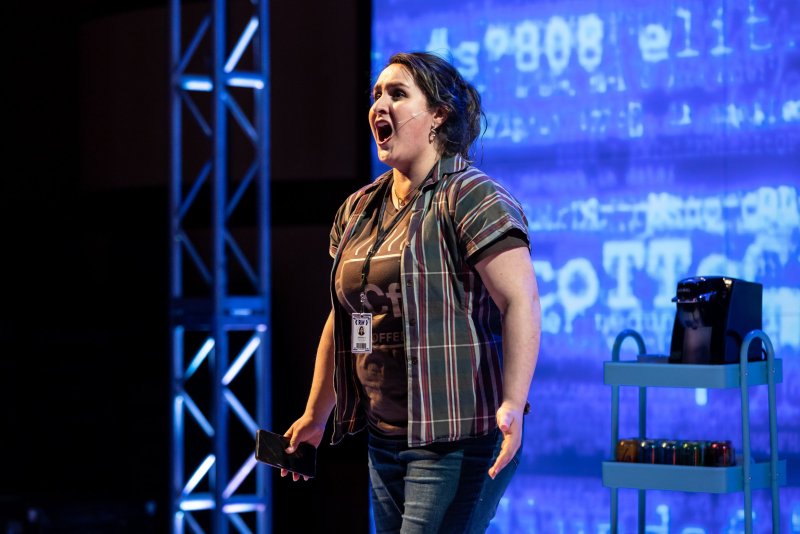 Onstage, a woman with dark hair sings, holding a phone. She wears jeans and a plaid shirt. Behind her, a blue screen displays blurred text, and a metal cart holds a coffee maker and cans.