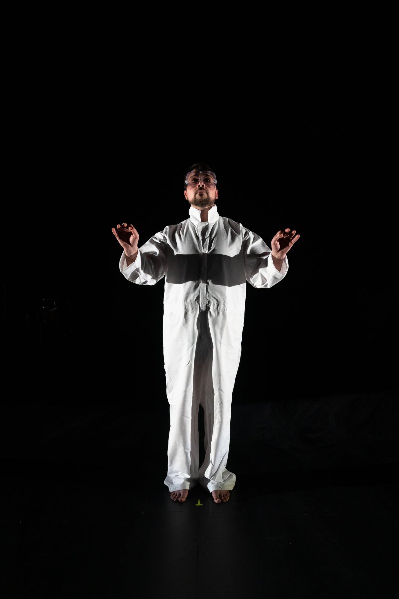 Man in white jumpsuit and goggles, arms raised, standing on a dark stage with a spotlight on him.