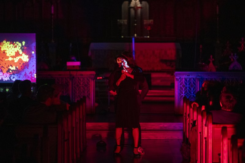 Performer in black dress holding microphone, bathed in red light, on stage with abstract projection, audience in pews.
