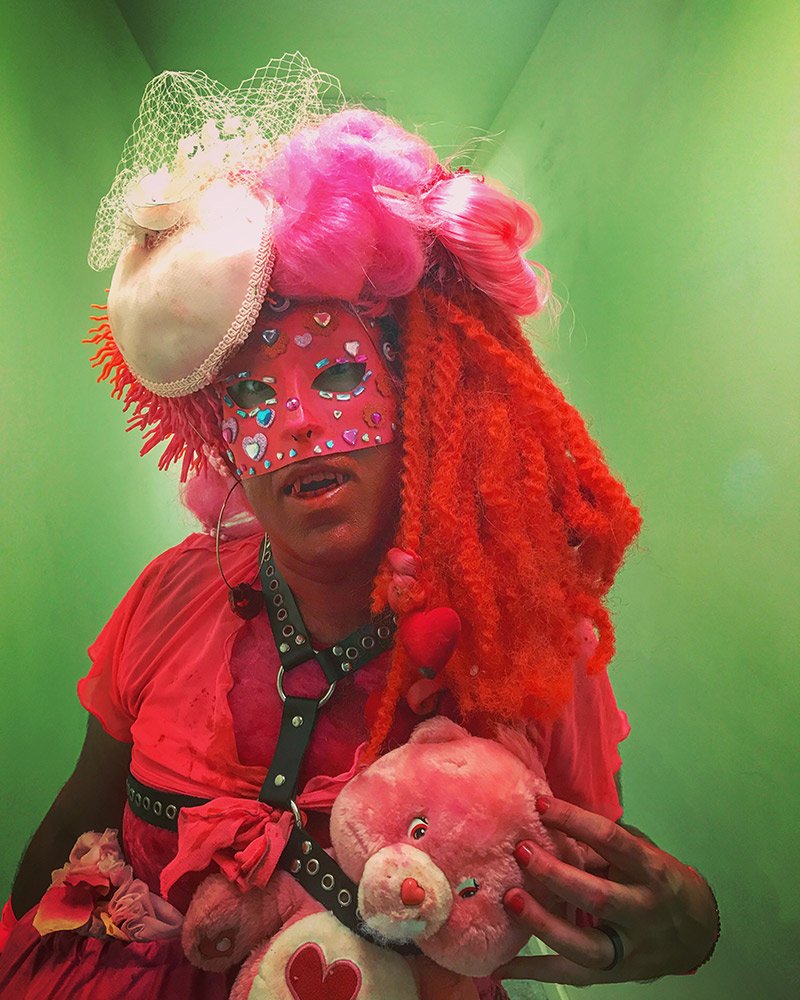 Person in pink wig and dress, red face mask, holding a pink Care Bear against a green background.