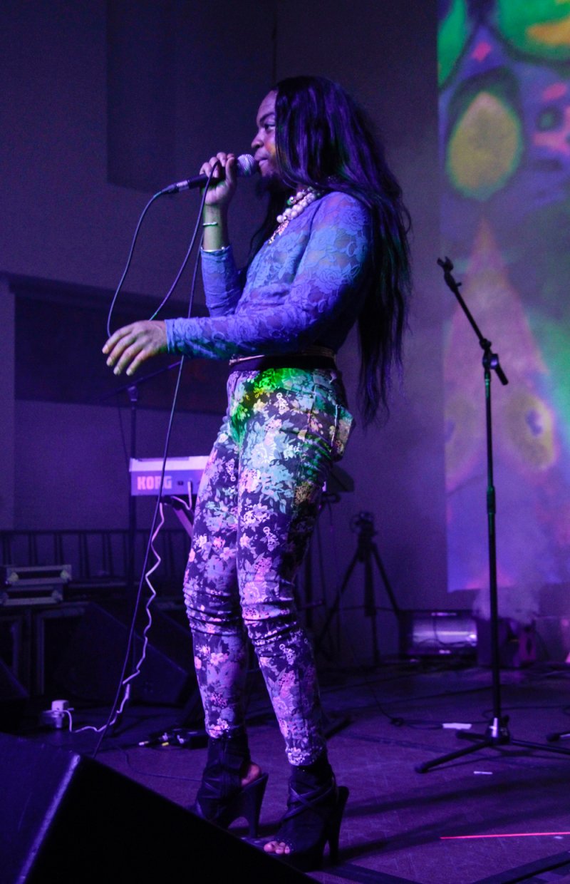 Performer sings on stage in purple light. She wears floral pants and a blue lace top.