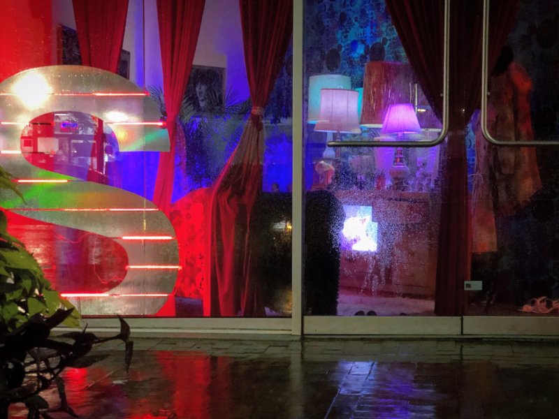 A storefront window reflecting a rainy street, revealing red curtains, neon lights, and glowing lamps inside.