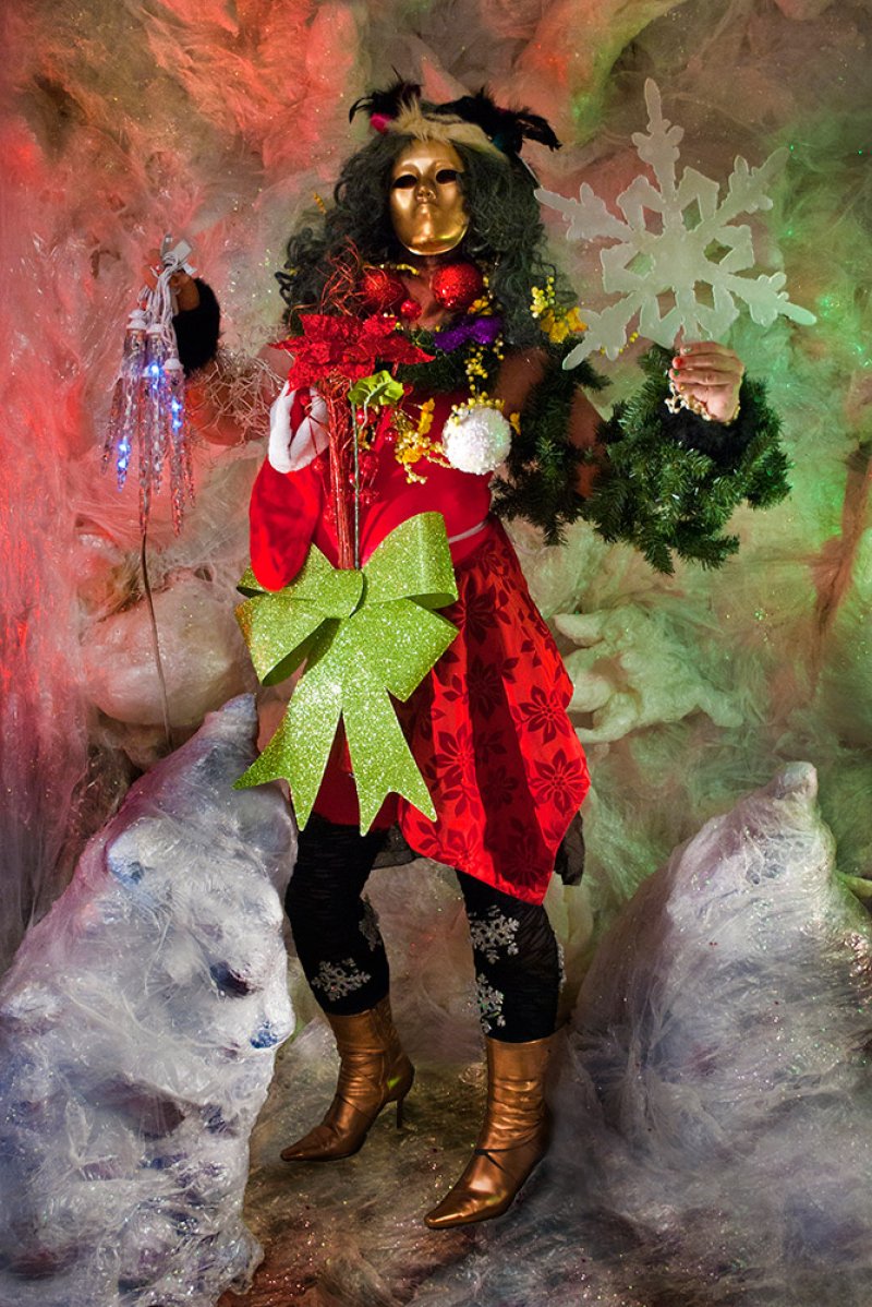 Full-body self-portrait of artist in a gold mask, red dress, and festive holiday decorations, holding a snowflake.