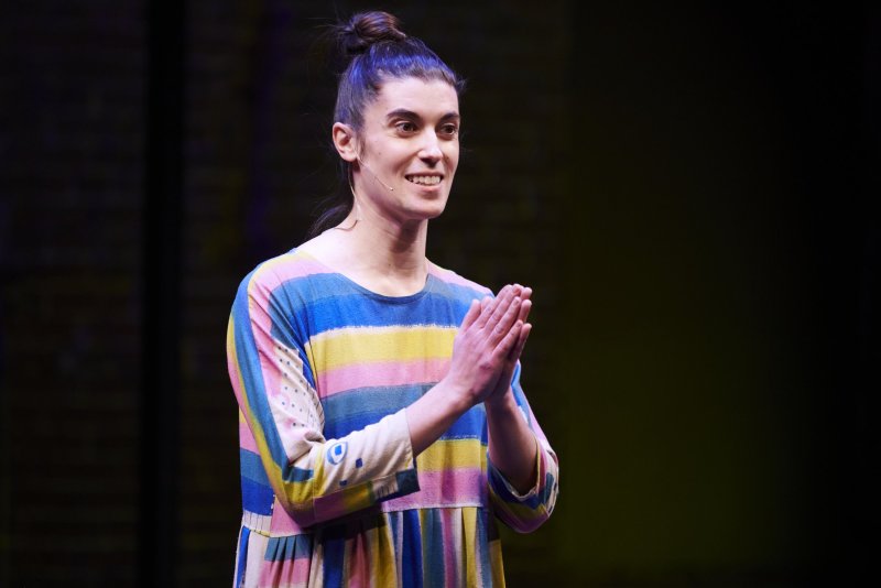 A smiling person with hair in a bun stands with hands pressed together. They wear a colorful striped dress with blue, yellow, and pink hues. The background is dark.