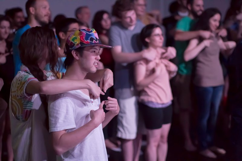 A diverse group of people in a dark room, some with arms around each other, looking forward intently.