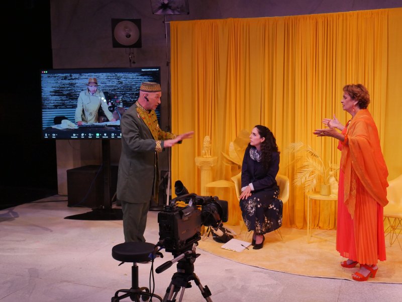 Stage production with three actors: one on a screen, two on stage with yellow curtains and props.