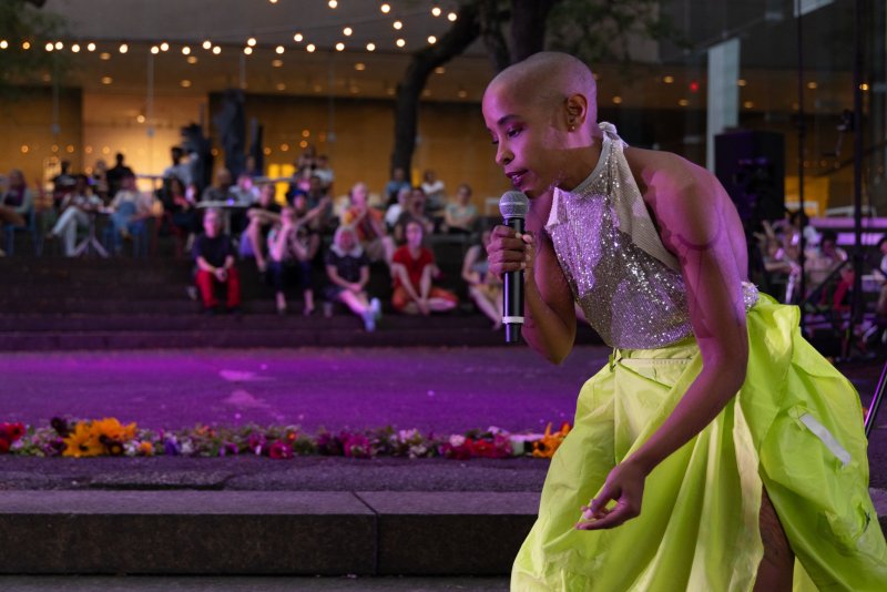 Performance shot: Bald person in sequined top and lime skirt sings into microphone on stage, audience in background. Purple stage lighting, flowers on stage edge.