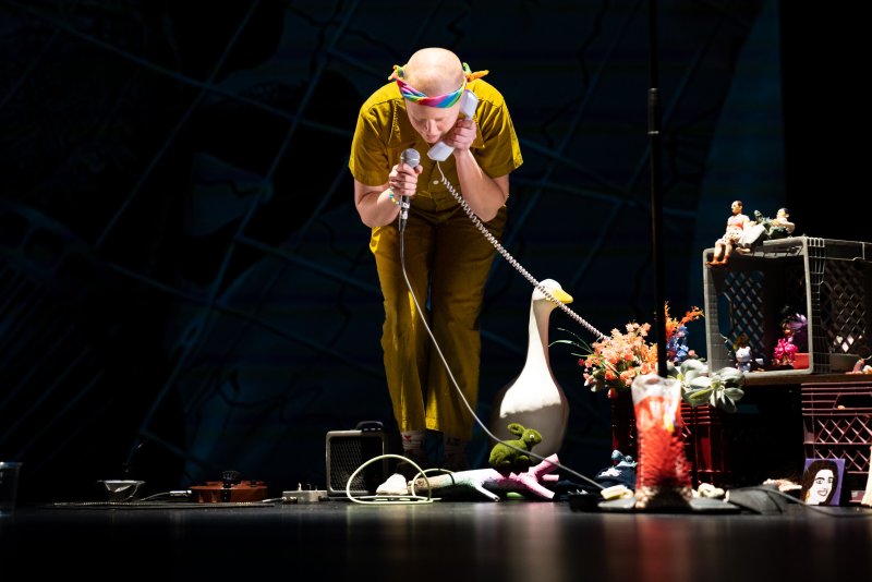 Performance art scene: Bald person in yellow jumpsuit holds a microphone and phone receiver. Stage cluttered with toys, flowers, a goose statue, and miniature figures on stacked crates. Dark background.