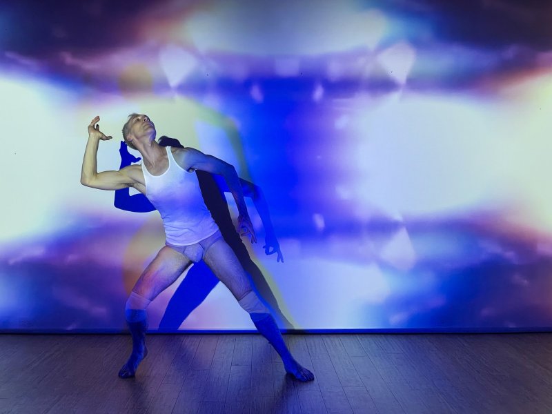 A male dancer in a white tank top and briefs poses against a vibrant blue and purple projected light background.