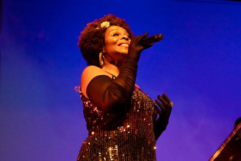 A Black woman with an afro, wearing a sequined dress and long black gloves, sings into a microphone against a blue background.