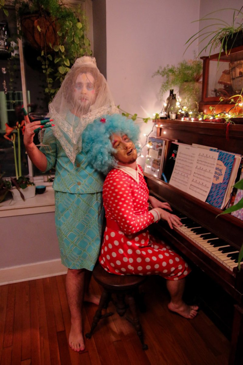 Two people in drag, one in a blue wig playing piano, the other in a veil and teal dress, both with painted faces.