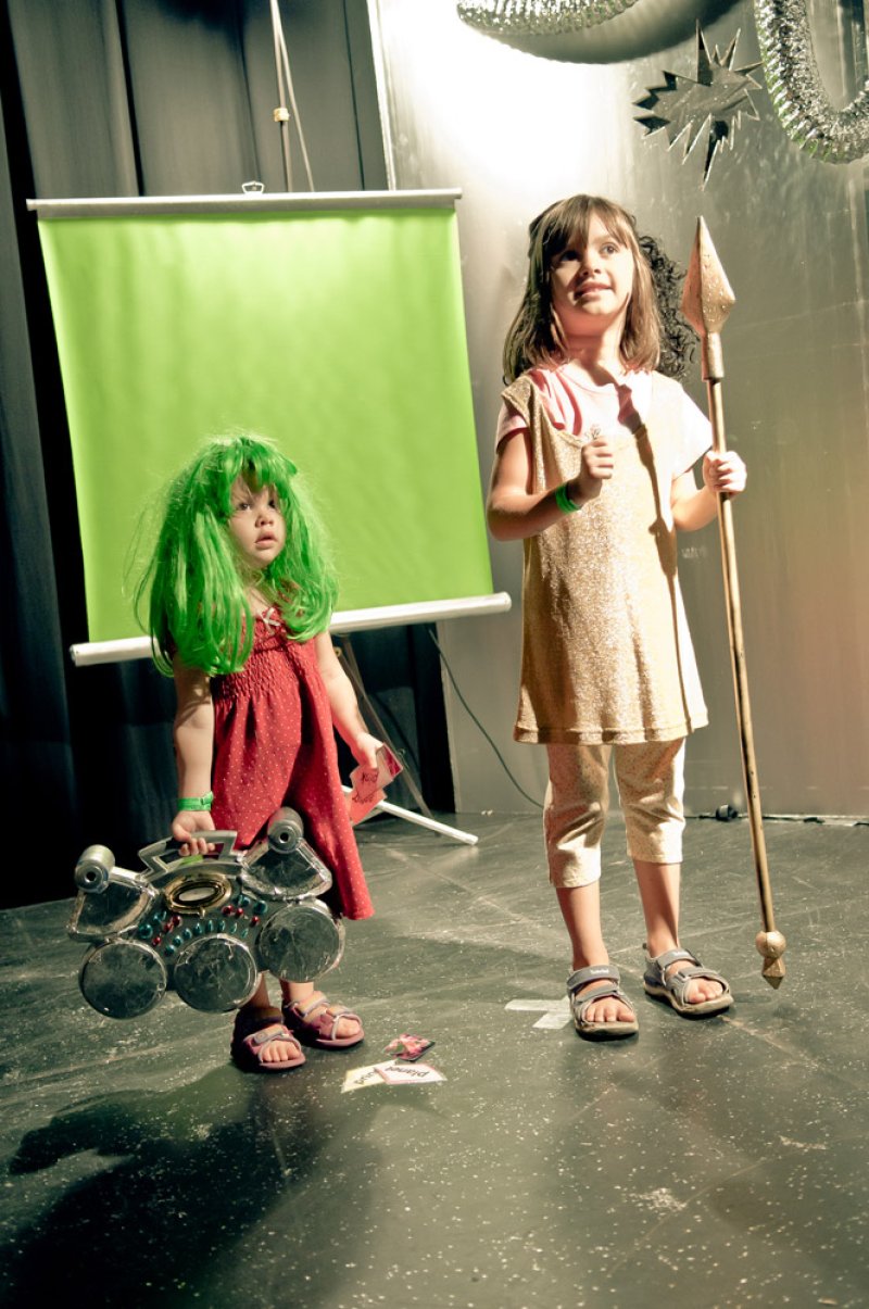 Two children in costumes stand on a stage with a green screen. One holds a toy drum set and wears a green wig, the other holds a spear. Tinsel decorations hang above.