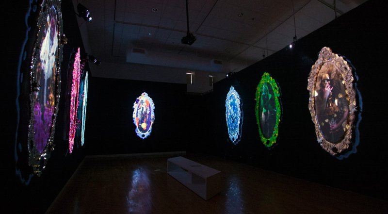 Dark room with six oval projections framed like mirrors, displaying figures in purple, pink, blue, green, and gold hues.