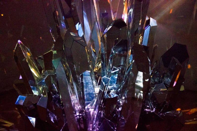 Sculptural installation of jagged, reflective crystal-like forms in dark tones, illuminated by purple and orange light.