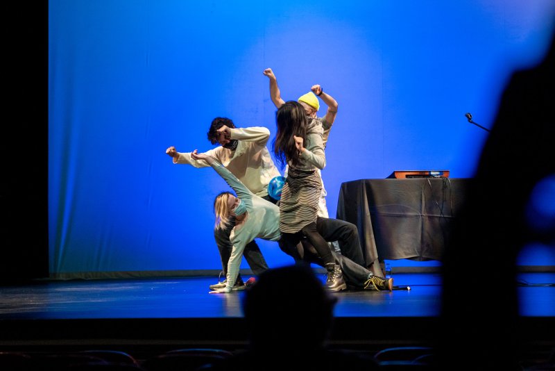 Four dancers in various poses on a stage with a bright blue background, one in foreground is blurry.