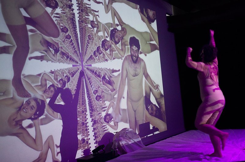 Performance with a dancer in a nude bodysuit facing a large projection of repeating figures and a central bearded man.