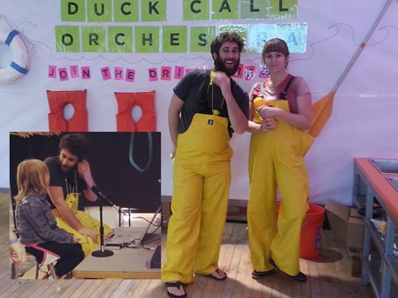 Two people in yellow waders stand in a room decorated with "Duck Call Orchestra" banners and nautical items.
