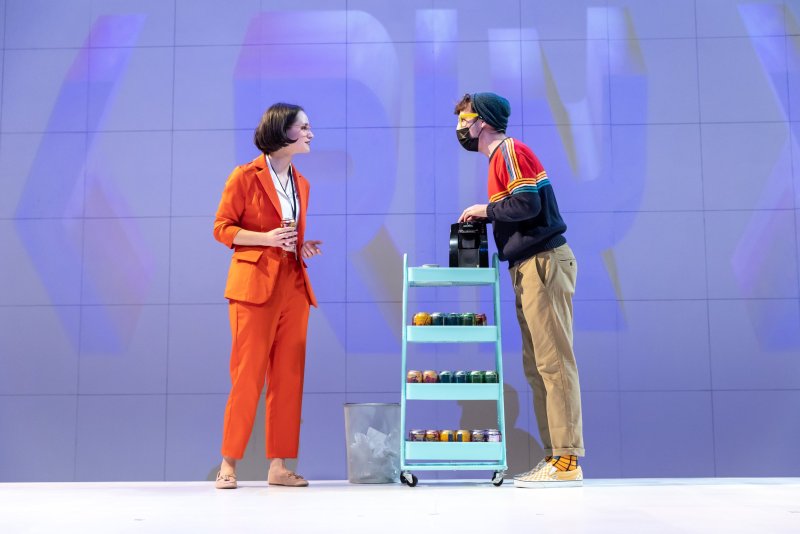 Two figures converse near a light blue cart stocked with colorful cans. A woman in an orange suit faces a person in a beanie and colorful sweater. The background is a light blue wall with a grid pattern.