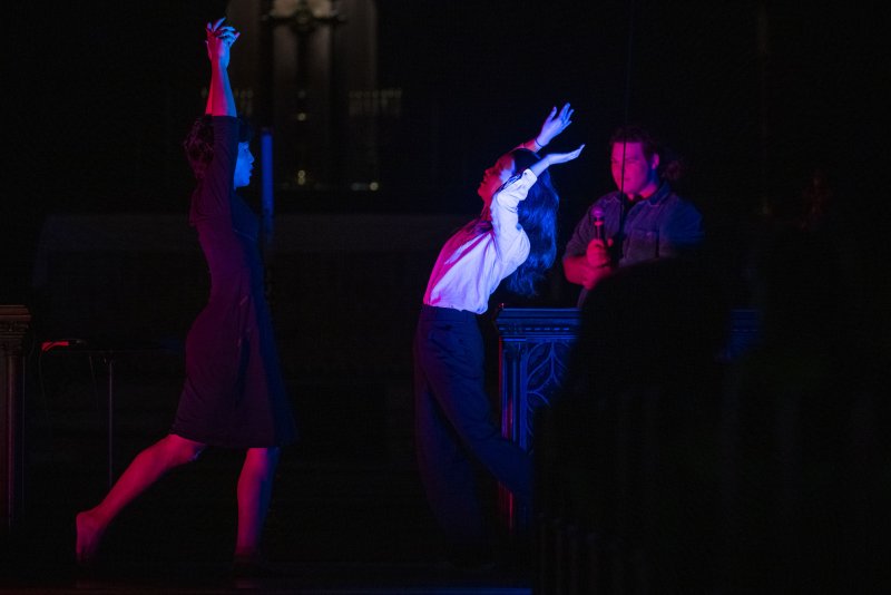 Two dancers in a dark space, illuminated by red and blue light, with a musician in the background.