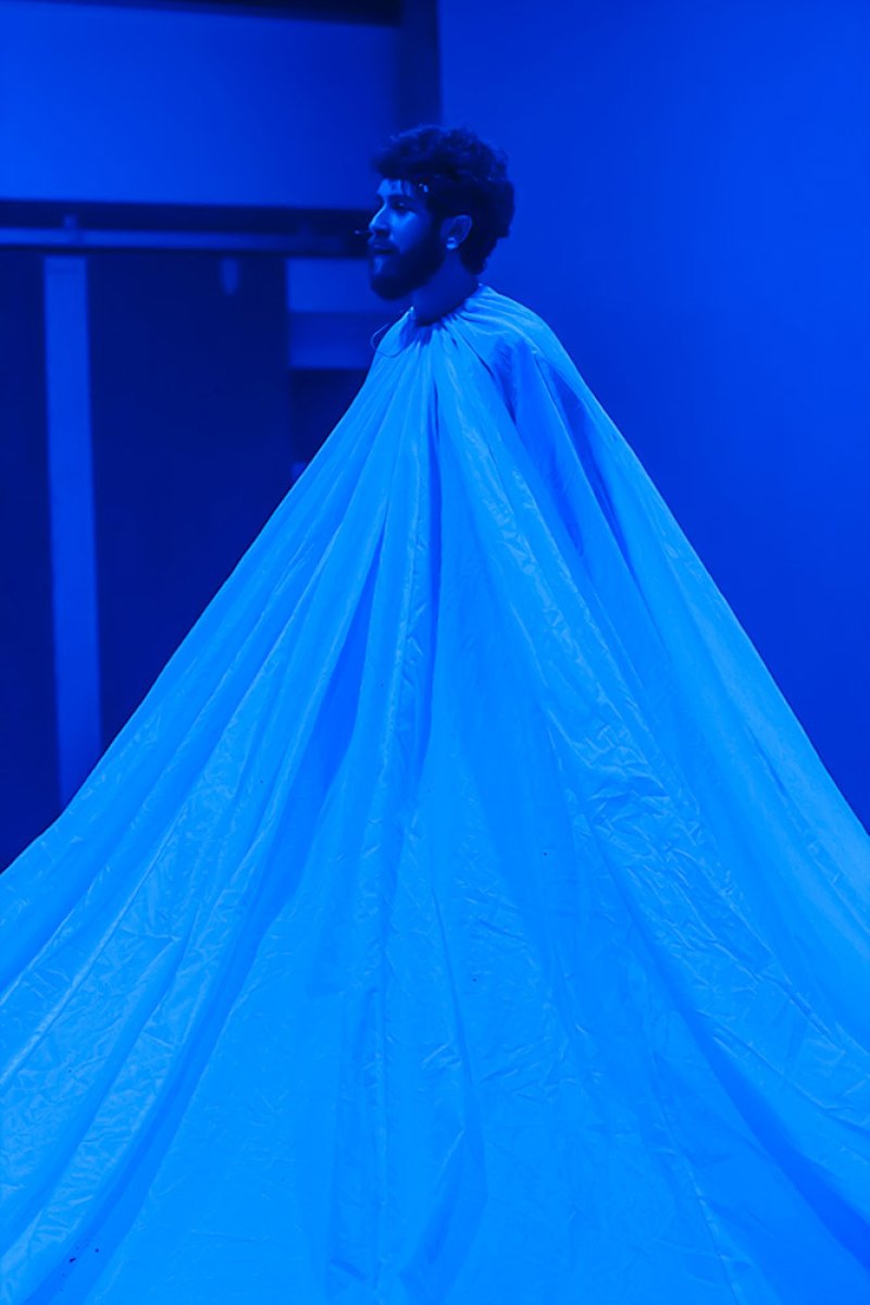 Man with beard and curly hair wearing a large, conical blue fabric garment in a dark, blue-lit room.
