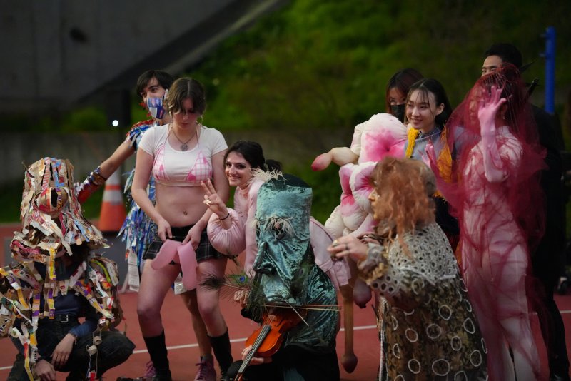 Performance art with people in eclectic, colorful costumes, including a figure in a teal mask playing a violin.