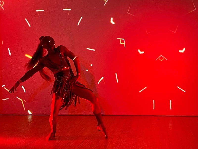 Dancer in a sparkling costume with long hair, striking a pose on a stage bathed in red light with abstract white lines projected around them.