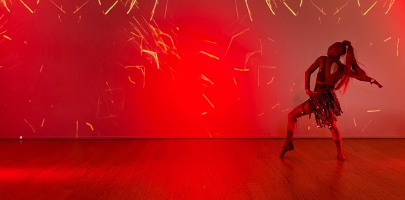 Dancer in a sequined costume striking a pose on a red-lit stage, with abstract golden light streaks projected onto the background.